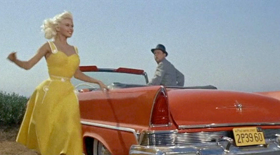 jayne with the full "dress on". she is on the left side of the photo (or film still) and we can see on the right a red coloured cabriolet car with a guy innit.